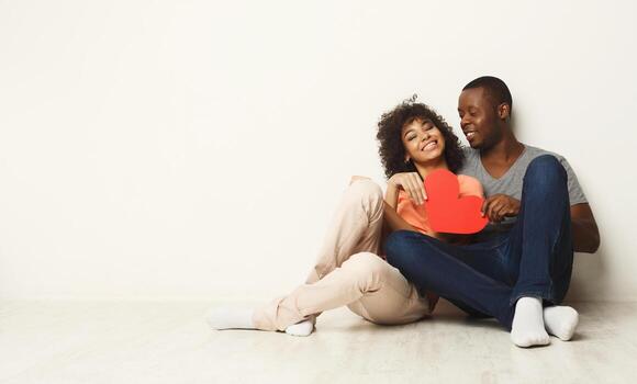 A joyful couple sits on the floor, smiling at each other while holding a red heart cutout. They are relaxed, showcasing love and connection in a minimalist setting. photo